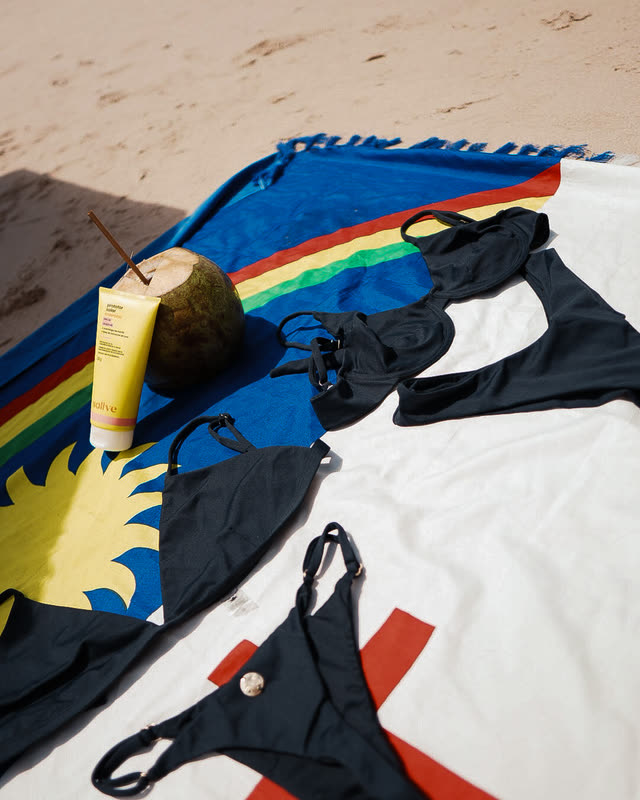 Biquínis da Sol Que Me Sirva sobre uma canga com a bandeira de Pernambuco, ao lado de um coco e um protetor solar